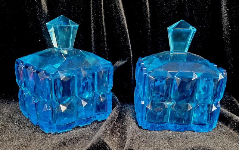 May include: Two matching, faceted, blue glass trinket boxes with geometric lids. The boxes have a square shape with multiple facets, creating a sparkling effect. The lids are also faceted and have a diamond-like shape.