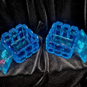 May include: Two blue, faceted, jewel-toned boxes with hinged lids. The boxes are open, revealing the interior. The boxes are made of a transparent material and sit on a dark, textured surface.