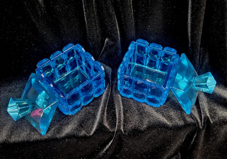 May include: Two blue, faceted, jewel-toned boxes with hinged lids. The boxes are open, revealing the interior. The boxes are made of a transparent material and sit on a dark, textured surface.