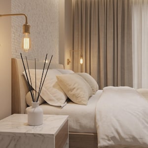 May include: A brass wall-mounted swing arm lamp with an exposed filament bulb, positioned above a white marble nightstand holding a white ceramic reed diffuser with black reeds, set beside a bed with white bedding and beige pillows.