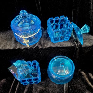 May include: Four pieces of blue glass decor are arranged on a dark surface. The items include a lidded jar with a cross charm, a square box, and a small round jar with a lid. The glass has a faceted design.