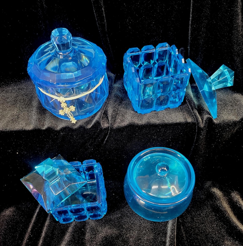 May include: Four pieces of blue glass decor are arranged on a dark surface. The items include a lidded jar with a cross charm, a square box, and a small round jar with a lid. The glass has a faceted design.
