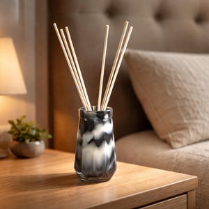 May include: A decorative reed diffuser with a black and white geometric pattern. The glass vessel holds several light-colored wooden reeds. The diffuser sits on a wooden nightstand next to a lamp and a small potted plant.