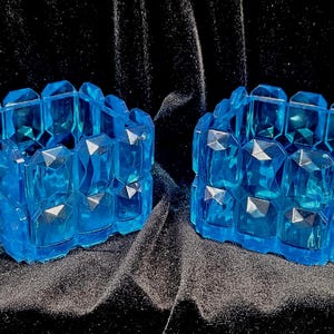 May include: Two matching, open, light blue, faceted, crystal-like boxes with lids. The boxes are on a black background. The lids are open, revealing the interior of the boxes. The boxes have a geometric design.