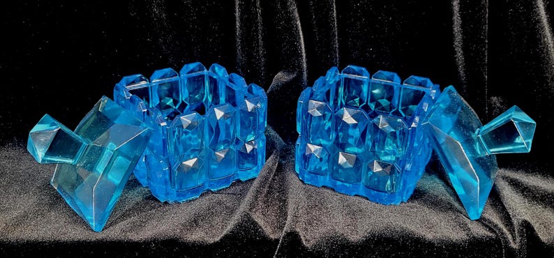 May include: Two matching, open, light blue, faceted, crystal-like boxes with lids. The boxes are on a black background. The lids are open, revealing the interior of the boxes. The boxes have a geometric design.