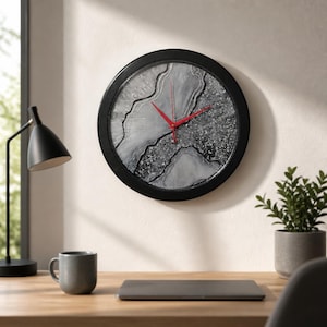 May include: A round wall clock with a black frame. The clock face features a gray and white abstract design with red hands. The clock is hanging on a light-colored wall.
