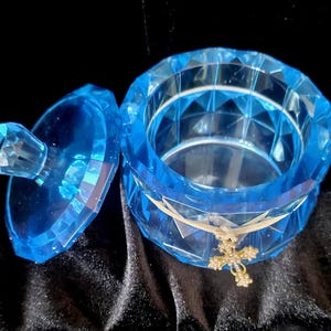 May include: A faceted, light blue trinket box with a matching lid. The open box has a gold-colored cross charm tied around it with a ribbon. The box is on a black, textured surface.