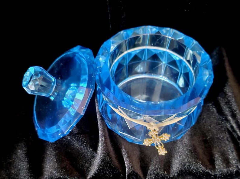 May include: A faceted, light blue trinket box with a matching lid. The open box has a gold-colored cross charm tied around it with a ribbon. The box is on a black, textured surface.