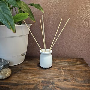 May include: A white ceramic essential oil diffuser with several wooden reeds. The diffuser sits on a dark wooden surface, next to a potted plant and decorative stones. The background is a textured, muted purple wall.