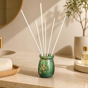 May include: A green reed diffuser vessel with gold-colored flecks and a smooth, rounded shape. Six thin, white, straight diffuser reeds are inserted into the top opening of the container, extending upwards in a fan-like arrangement.