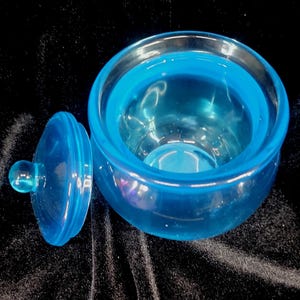 May include: A transparent, light blue glass jar with a matching lid. The jar is round with a wide opening, and the lid has a small, round handle. The jar and lid are made of a smooth, glossy glass material.