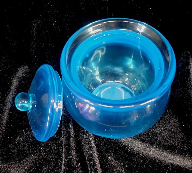 May include: A transparent, light blue glass jar with a matching lid. The jar is round with a wide opening, and the lid has a small, round handle. The jar and lid are made of a smooth, glossy glass material.