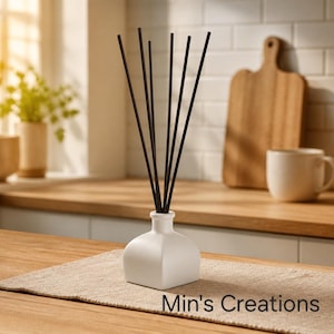 May include: A white square-based ceramic reed diffuser bottle with several thin black reeds extending upwards. The text Min's Creations is printed in black on the lower right side of the image. The diffuser is placed on a light tan fabric mat.