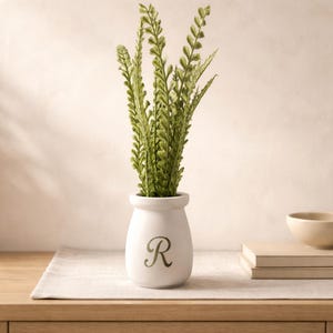 May include: A white ceramic vase with a green letter "R" holds several green leafy stems. The vase sits on a wooden table with a neutral-colored placemat. A small bowl and books are in the background.