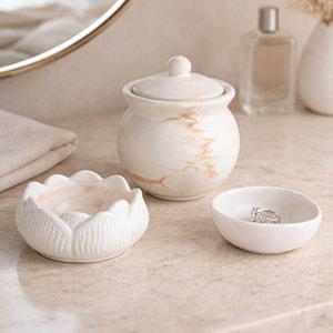 May include: A collection of white ceramic bathroom accessories. The set includes a small lidded jar with a marble effect, a lotus-shaped dish, and a small round dish holding a ring. The items are arranged on a light-colored countertop.