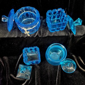 May include: A collection of translucent blue decorative boxes and containers. The set includes round and square shapes, some with lids. One container has a small cross charm. The items are arranged on a dark, velvety surface.