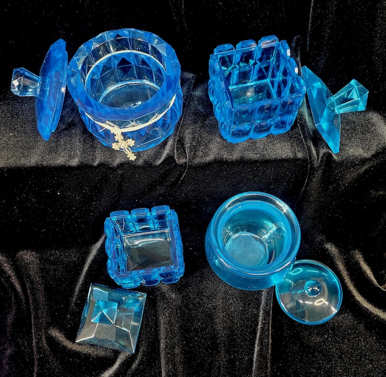 May include: A collection of translucent blue decorative boxes and containers. The set includes round and square shapes, some with lids. One container has a small cross charm. The items are arranged on a dark, velvety surface.
