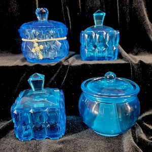 May include: Four blue glass jars with lids. The jars have different shapes and designs, including faceted and rounded styles. One jar is adorned with a gold-colored cross charm and a cream-colored band. The jars are displayed against a dark background.