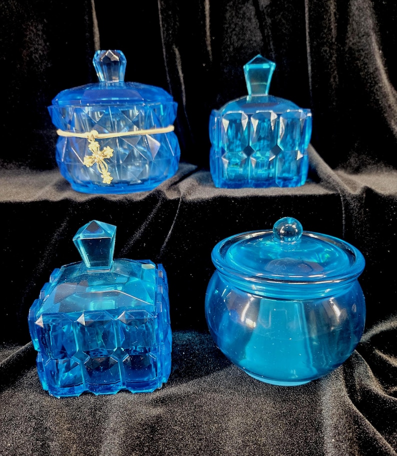 May include: Four blue glass jars with lids. The jars have different shapes and designs, including faceted and rounded styles. One jar is adorned with a gold-colored cross charm and a cream-colored band. The jars are displayed against a dark background.