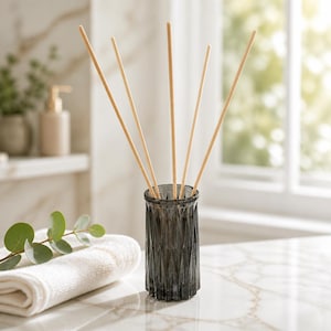 May include: A dark gray, faceted glass reed diffuser vessel with a cylindrical shape and a textured geometric pattern. Five light-colored, slender wooden reeds are inserted into the opening, extending upward in a fan-like arrangement.