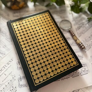 Op de afbeelding: Een vintage boek met een zwarte kaft en een gouden stippenpatroon. Het boek ligt op bladmuziek, met een vergrootglas in de buurt. De kaft heeft een gouden rand en een patroon van gouden cirkels.