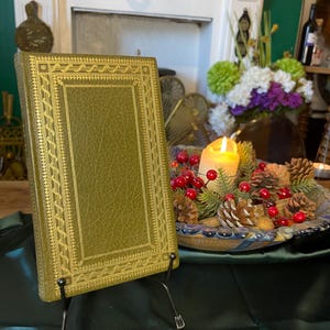 May include: An antique book with a green cover and gold embossed detailing, displayed on a black metal stand. The book is the focal point, set against a backdrop of a fireplace, flowers, and a candlelit centerpiece.