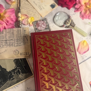 Op de afbeelding: Een rood notitieboek met een gouden geometrisch patroon, een roze lint-bladwijzer en een verzameling vintage ephemera. De items omvatten oude brieven, een vergrootglas, een briefopener en gedroogde rozenblaadjes.