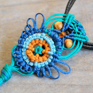 Pode incluir: Um colar de macram&eacute; azul, verde e laranja com contas de madeira e um tassel. O colar &eacute; feito com uma t&eacute;cnica de n&oacute;.