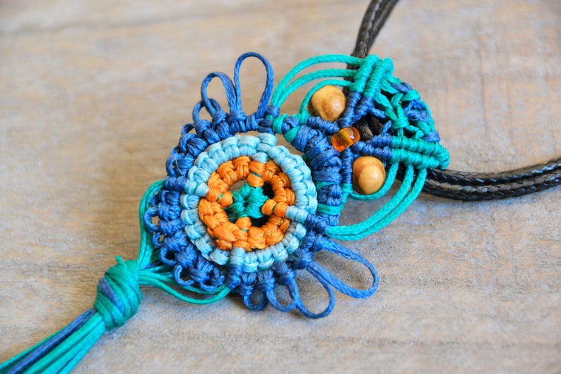 Pode incluir: Um colar de macram&eacute; azul, verde e laranja com contas de madeira e um tassel. O colar &eacute; feito com uma t&eacute;cnica de n&oacute;.