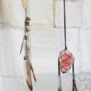 May include: Two crystal dreamcatchers hanging from a white fabric background. The dreamcatcher on the left has a clear crystal with a brown and white feather. The dreamcatcher on the right has a pink crystal with a brown and white feather.
