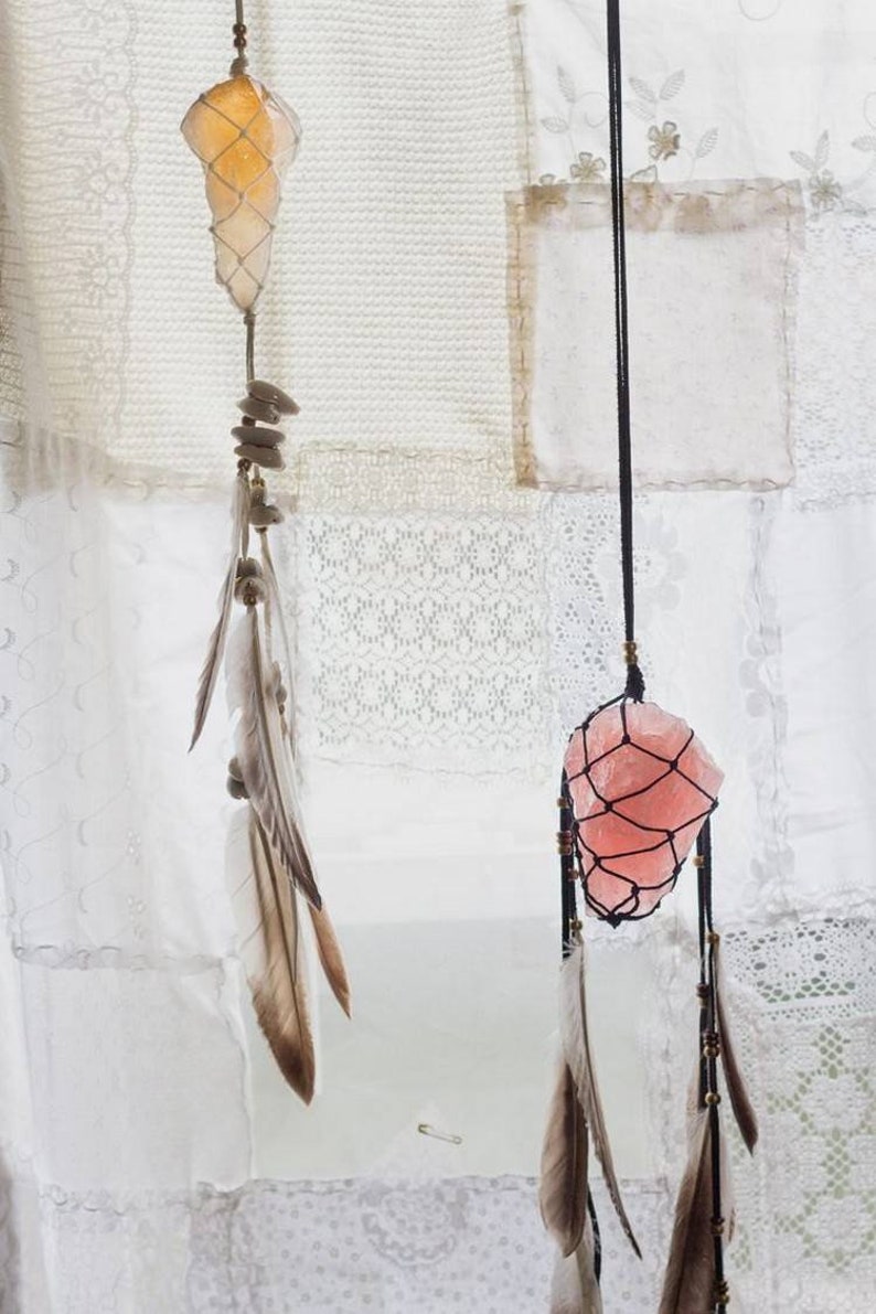 May include: Two crystal dreamcatchers hanging from a white fabric background. The dreamcatcher on the left has a clear crystal with a brown and white feather. The dreamcatcher on the right has a pink crystal with a brown and white feather.