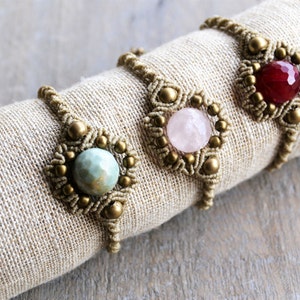 Puede incluir: Tres pulseras de macram&eacute; con cuentas de piedras preciosas de diferentes colores. Las pulseras est&aacute;n hechas con cord&oacute;n marr&oacute;n y cuentas doradas. Las piedras preciosas son de color verde claro, rosa y rojo.