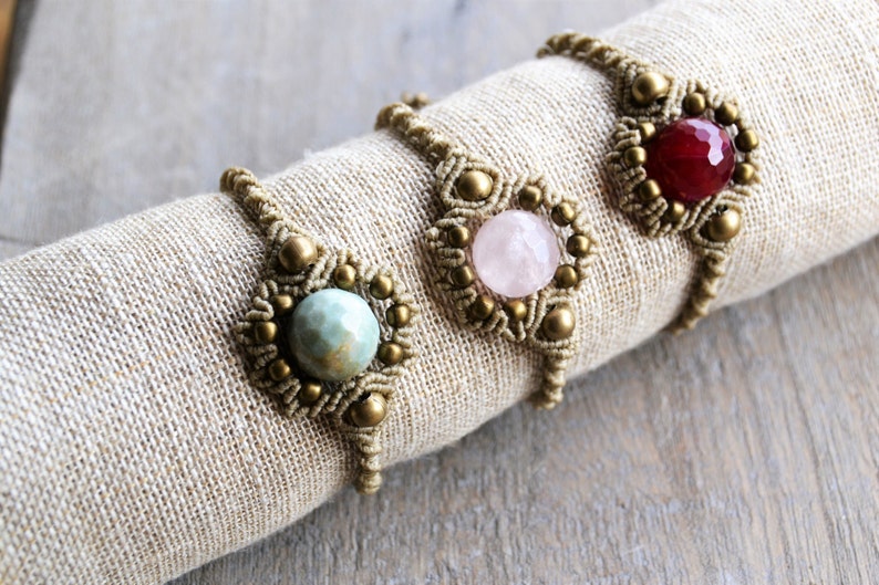 Puede incluir: Tres pulseras de macram&eacute; con cuentas de piedras preciosas de diferentes colores. Las pulseras est&aacute;n hechas con cord&oacute;n marr&oacute;n y cuentas doradas. Las piedras preciosas son de color verde claro, rosa y rojo.
