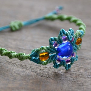 Puede incluir: Un brazalete de macram&eacute; verde, azul y morado con una cuenta de vidrio azul y cuentas de vidrio naranja.