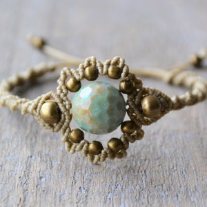 Puede incluir: Un brazalete de macram&eacute; con una cuenta de piedra preciosa verde y marr&oacute;n en el centro. El brazalete est&aacute; hecho con cord&oacute;n beige y cuentas doradas.