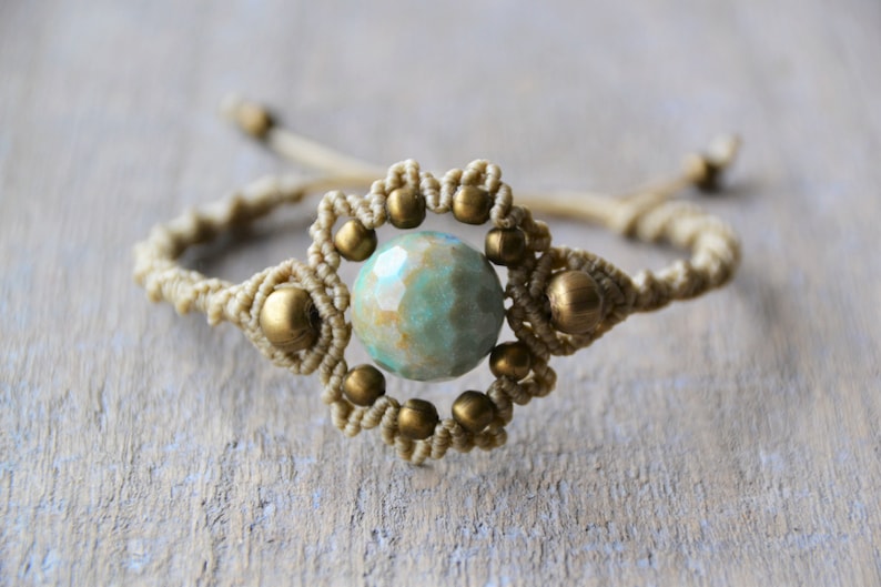 Puede incluir: Un brazalete de macram&eacute; con una cuenta de piedra preciosa verde y marr&oacute;n en el centro. El brazalete est&aacute; hecho con cord&oacute;n beige y cuentas doradas.
