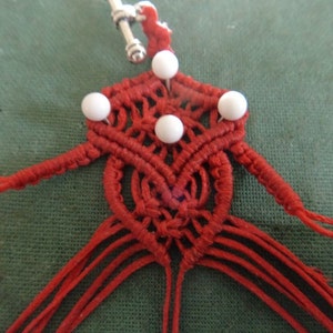 Puede incluir: Nudo de macram&eacute; rojo con cuentas blancas, un lazo plateado en la parte superior y largas cuerdas rojas que cuelgan.