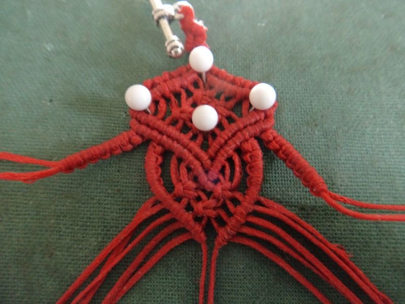Puede incluir: Nudo de macram&eacute; rojo con cuentas blancas, un lazo plateado en la parte superior y largas cuerdas rojas que cuelgan.