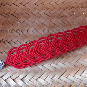 Puede incluir: Un brazalete de macram&eacute; rojo con un cierre plateado con dise&ntilde;o de rosa. El brazalete est&aacute; hecho con una serie de nudos interconectados que forman un patr&oacute;n de corazones.
