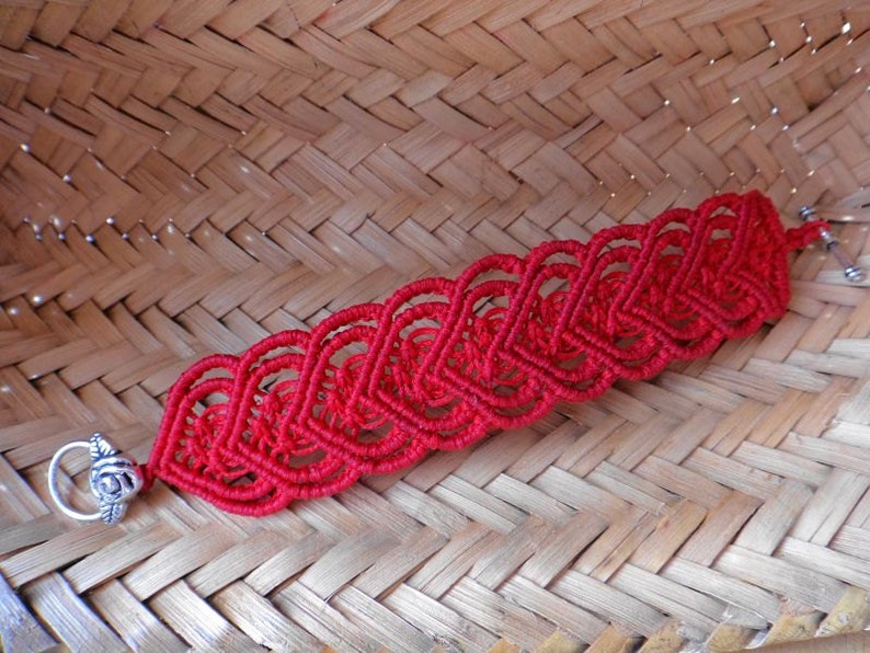 Puede incluir: Un brazalete de macram&eacute; rojo con un cierre plateado con dise&ntilde;o de rosa. El brazalete est&aacute; hecho con una serie de nudos interconectados que forman un patr&oacute;n de corazones.