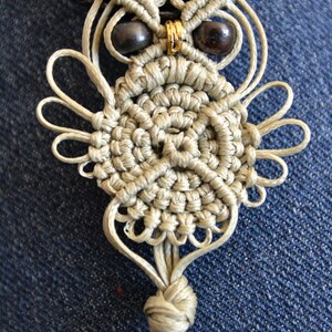 Pode incluir: Um pingente de coruja de macram&eacute; bege com contas pretas para os olhos e um detalhe dourado. A coruja est&aacute; presa a um cord&atilde;o de couro preto.