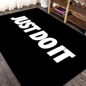 Könnte beinhalten: Schwarzer rechteckiger Teppich mit den Worten "JUST DO IT" in großen weißen Großbuchstaben. Der Teppich liegt auf einem dunklen Holzboden. Der Text ist über den Teppich abgewinkelt.