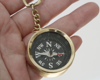 May include: A gold-colored compass keychain with a black face and white markings. The compass has a red needle and is attached to a gold-colored chain and a split ring.