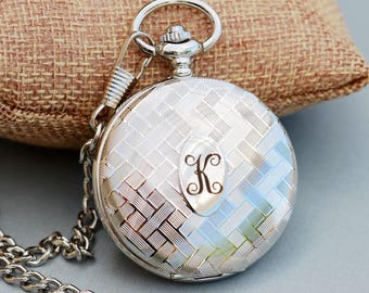 May include: A silver pocket watch with a chain and a clasp. The watch face has a patterned design and an engraved initial "K" in the center.