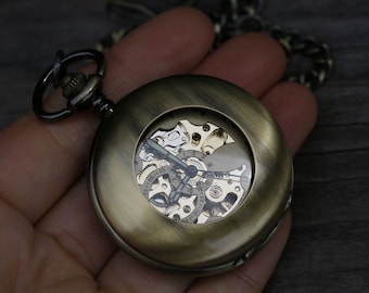May include: A gold-toned pocket watch with a skeletonized face, revealing the intricate inner workings of the watch. The watch has a black chain and a decorative loop.