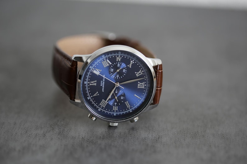 May include: A silver-toned wristwatch with a blue dial and brown leather band. The watch face features Roman numerals and three smaller dials. The band is a dark brown, textured leather. The watch is a classic accessory.