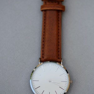 Puede incluir: Un reloj de pulsera plateado con una esfera blanca y una correa de cuero marr&oacute;n.