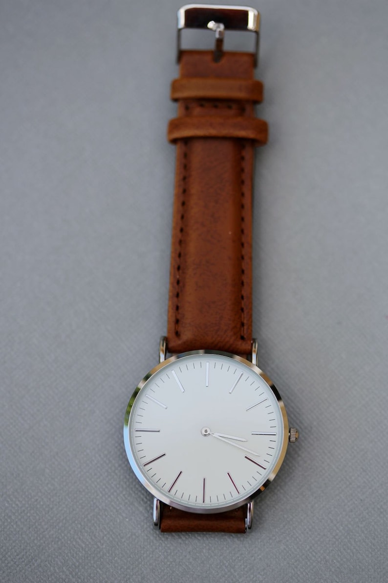Puede incluir: Un reloj de pulsera plateado con una esfera blanca y una correa de cuero marr&oacute;n.
