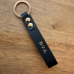 May include: Black leather keychain with gold-colored hardware. The keychain features a gold ring and two gold studs. The leather strap is embossed with the initials "D ♡ A" in gold lettering. The keychain is on a wooden surface.