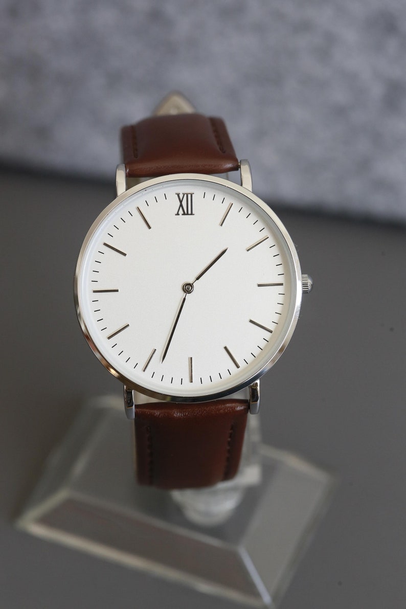 May include: A silver watch with a white face and brown leather band. The watch face has Roman numeral hour markers and black hands.